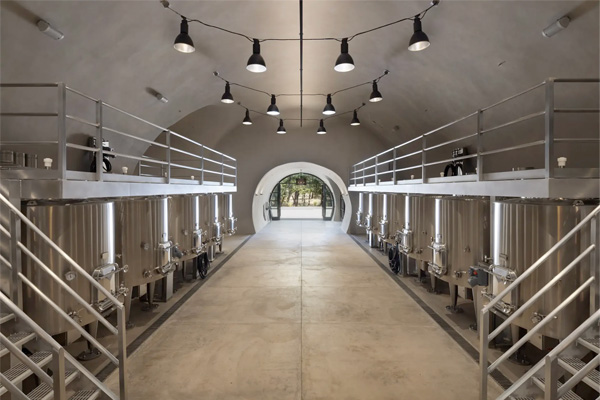 A modern brewery interior with stainless steel fermentation tanks lining both sides of a central walkway, illuminated by overhead lights.