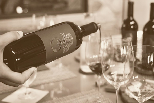 A hand pours red wine into a glass on a table set with additional wine glasses and wine bottles in the background. The image is in sepia tone.