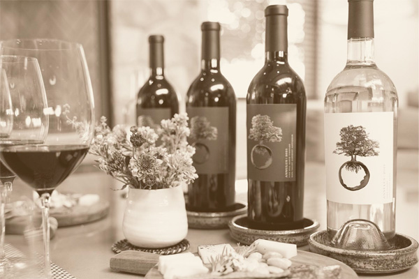 Four wine bottles, three red and one white, placed on a table with a wine glass, a small flower vase, and a serving board with snacks.