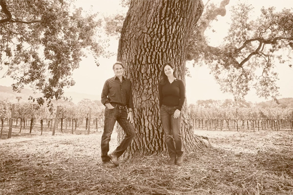 Two people stand beside a large tree in a vineyard. The vineyard stretches out in rows in the background, and the setting appears peaceful and natural.