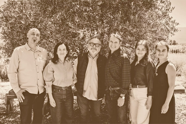 Six people stand together outdoors in front of a tree, posing for a group photo. The image is sepia-toned.