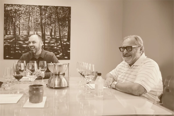 Two men are seated at a table with multiple wine glasses, seemingly engaged in a wine tasting. A photograph of trees hangs on the wall behind them.