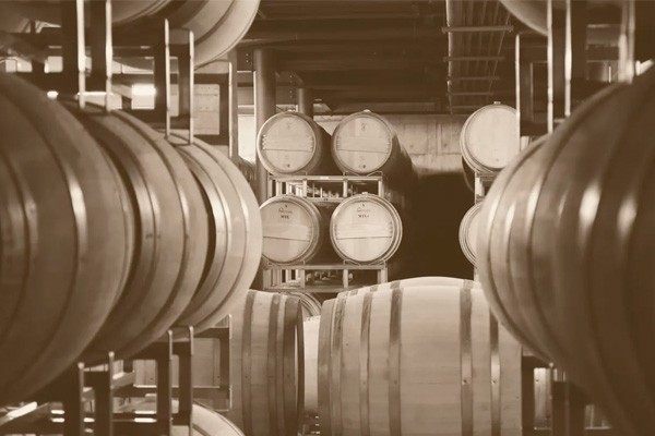 A sepia-toned image showing rows of stacked wooden barrels in a winery cellar.