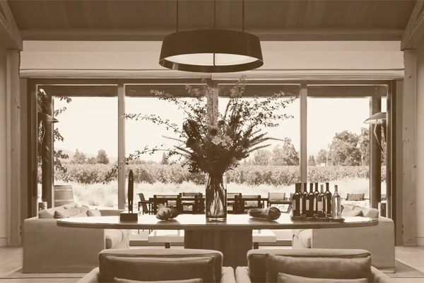 A spacious room with large windows showcasing an outdoor vineyard view. A central table is adorned with flowers, wine bottles, and decorations. Comfortable seating areas surround the table.