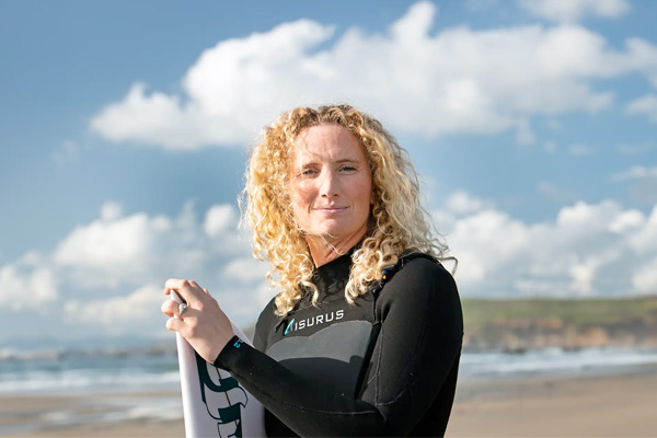 A person with curly blonde hair, wearing a wetsuit, stands on a beach holding a surfboard against a backdrop of clouds and ocean waves.