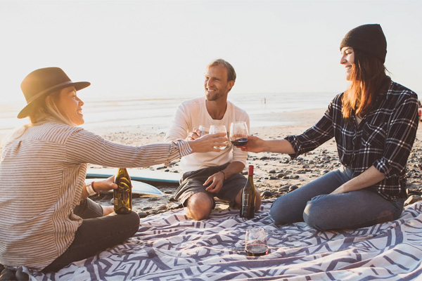 Three people sit on a beach blanket, toasting with drinks. A man is in the middle, and two women are on either side, one wearing a hat and the other a beanie. The ocean is visible in the background.