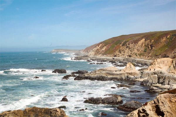 Rocky coastline with waves crashing against the shore under a clear blue sky. Rugged hills and cliffs stretch into the distance.