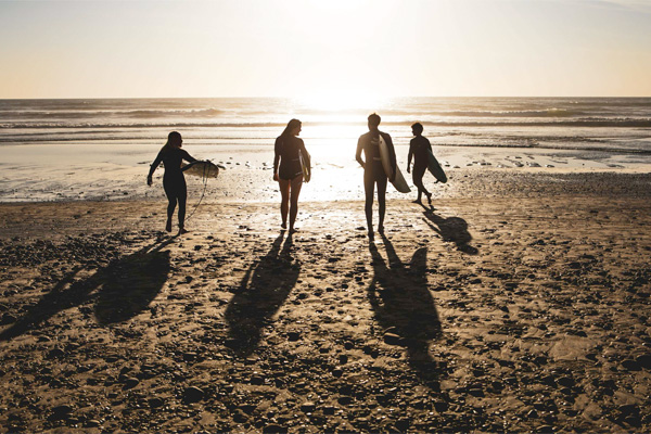 Four people in wetsuits walk along a beach at sunset, carrying surfboards and casting long shadows on the sand.