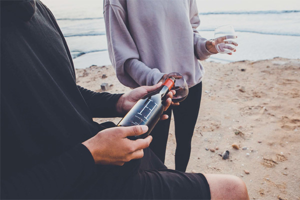 Two people at the beach, one holding a wine bottle and the other holding a wine glass.