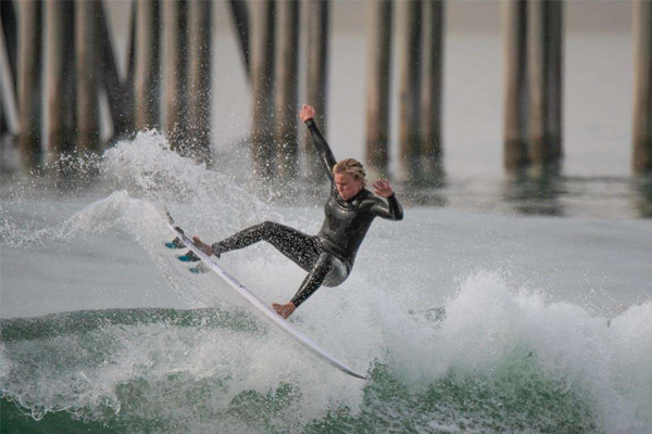 A person in a wetsuit performs an aerial maneuver on a surfboard above a wave near wooden pilings.