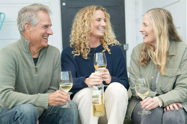 Three people sitting and laughing while holding glasses of white wine, with one person in the middle holding a wine bottle.