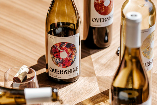 Four bottles of Overshine wine are arranged on a wooden surface, along with a cork and a partially filled wine glass. The label features circular artwork.