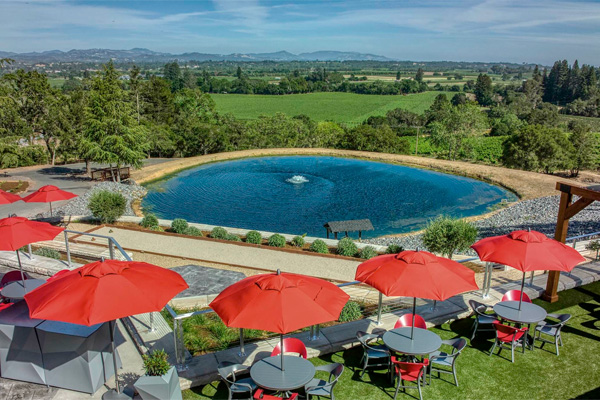 A scenic outdoor seating area with red umbrellas and tables overlooks a round pond and expansive green landscape under a blue sky.