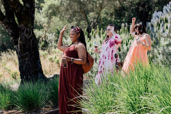 Three women walking outdoors, smiling and waving, with greenery and trees in the background.