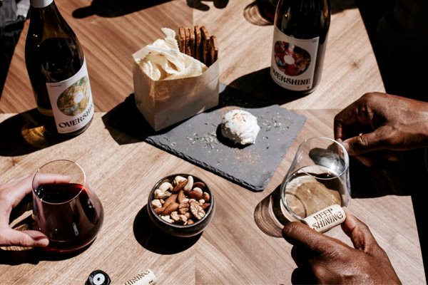 A wooden table with snacks, nuts, and cheese, surrounded by bottles of wine. Four hands are holding wine glasses and corks with 'Keep Shining' message.