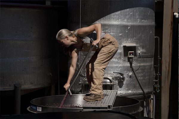 Jasmine Hirsch in work overalls uses a tool to stir fermenting wine inside a large metal tank in an industrial setting.