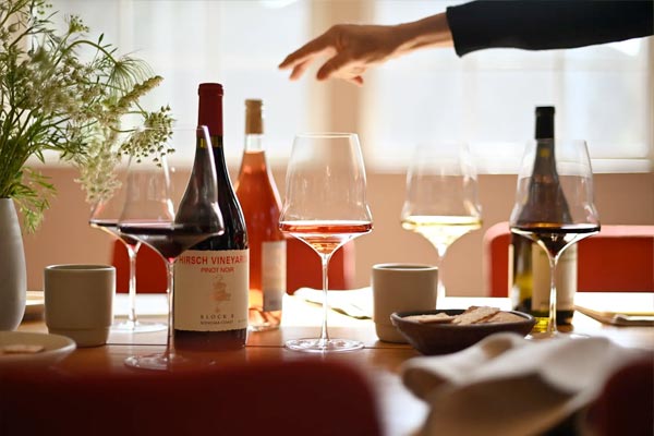 A table set with wine bottles, wine glasses, cups, a small plate of bread, and flowers, with a hand reaching toward the bottles.